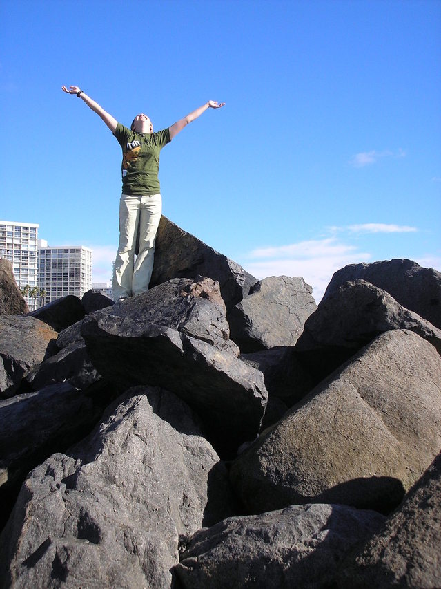 A photo of a happy or merry girl standing on large black rocks with her arms raised.