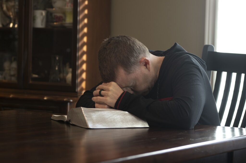 A man with his head down on folded arms against an open Bible on a table illustrating that the only way to understand God is through pray and fellowship with Him