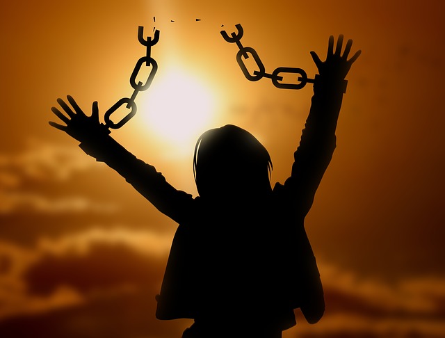 A image of a silhouette of a woman with her hands and shackled wrists up in the air waving a broken chain of being delivered against a hazy sun in a pale orange sky.