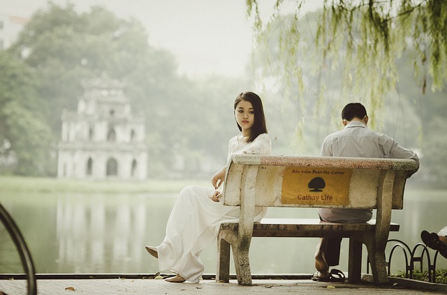 A young Vietnamese woman, sitting opposite a young man on a bench beside a calm lake, displaying an expression of emptiness.