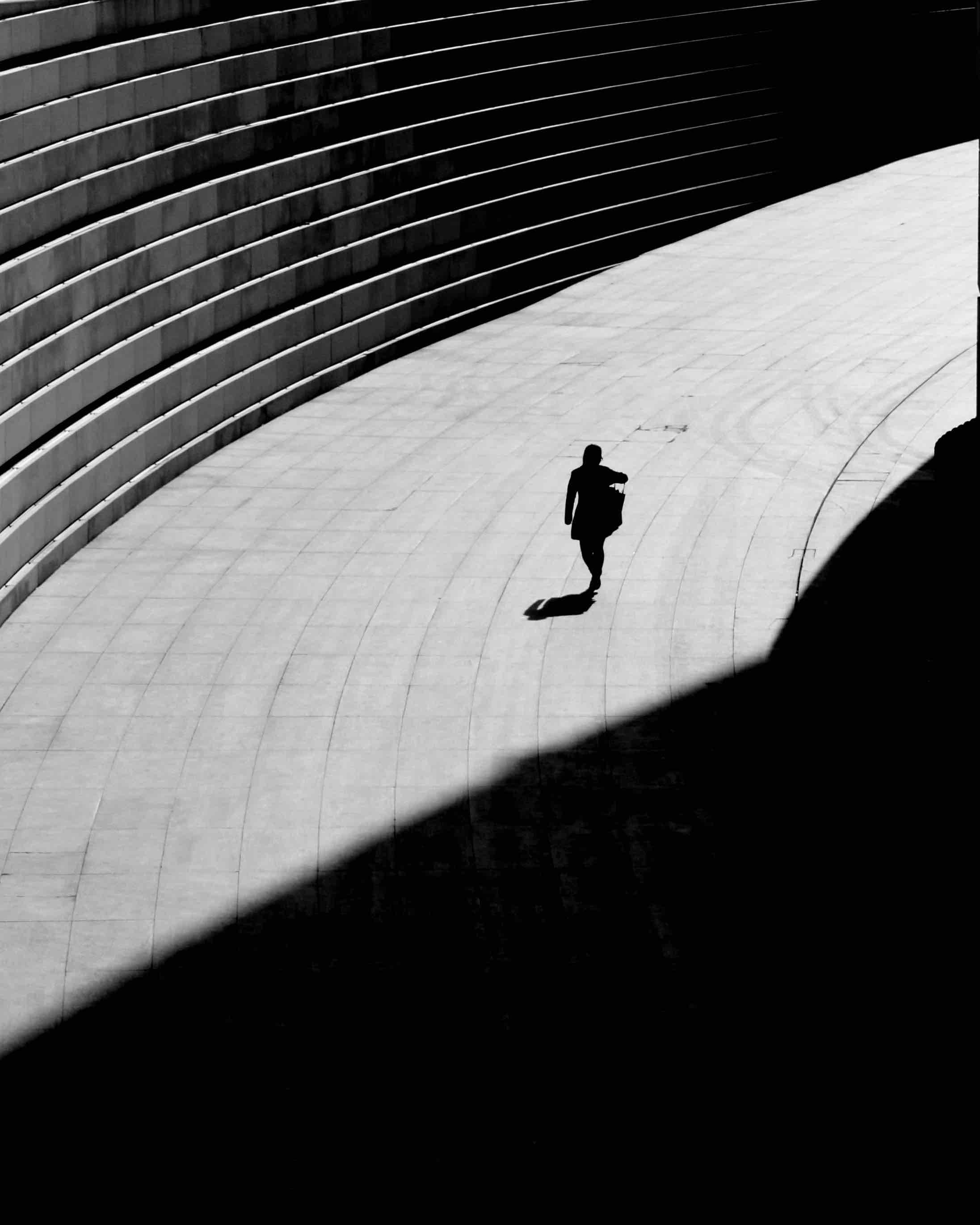 A silhouette of person walking on an open fully sun-lit pathway towards a shadow.
