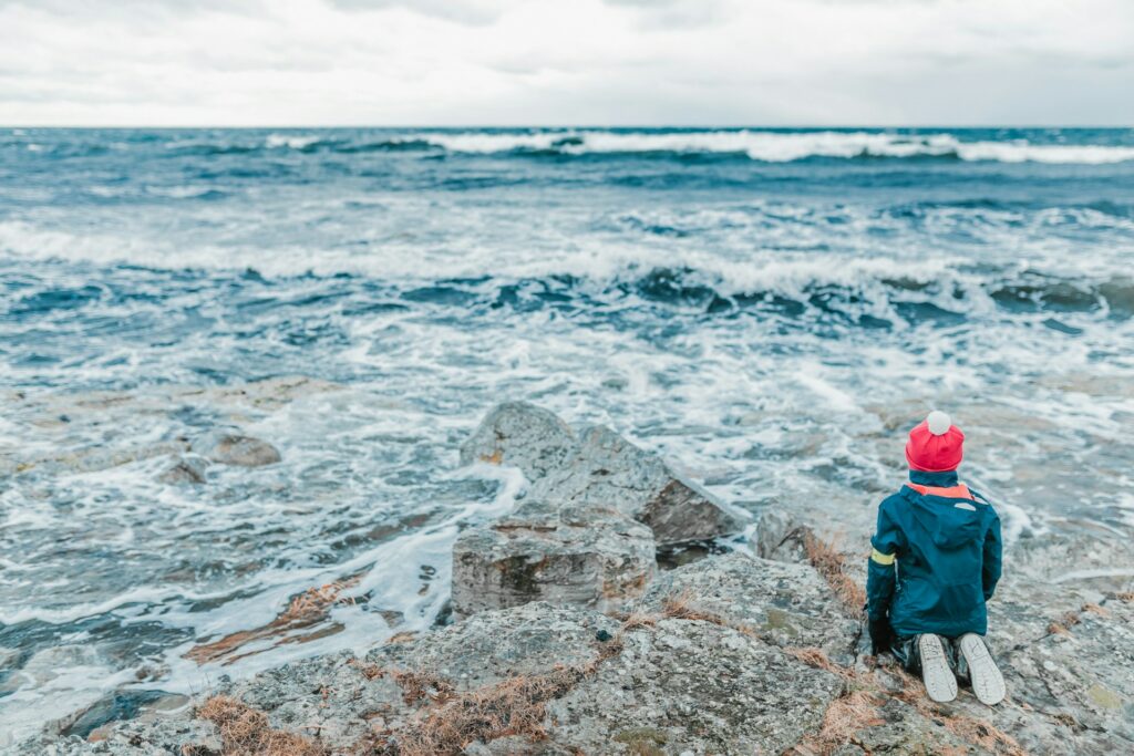 A child with coat and hat kneeling on rocks overlooking ocean surf, a reminder of being dependent on God in a difficult world