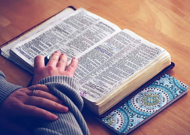 Open Bible laying flat on hardwood table with hands folded and resting partially near lower portion of book