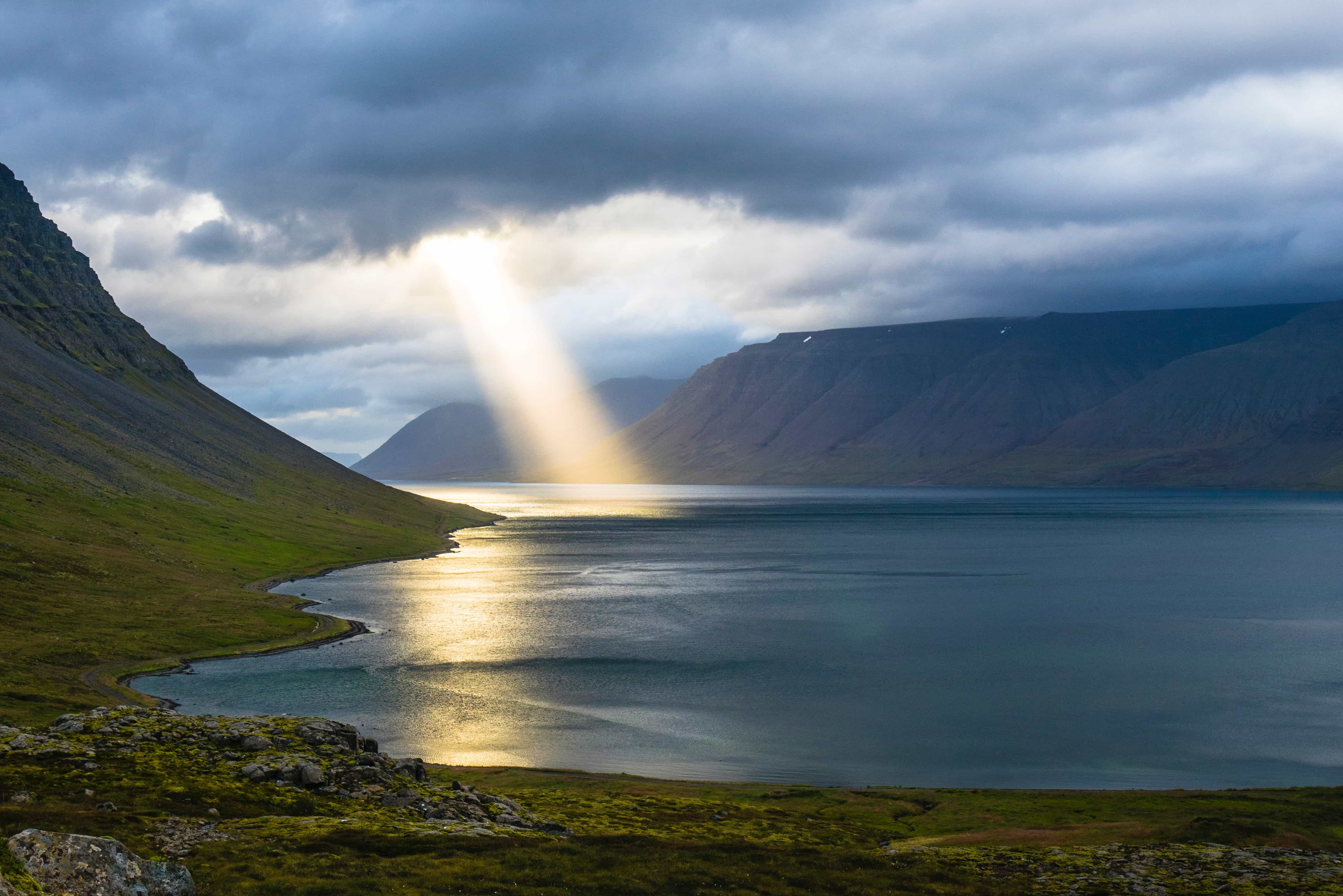 A beam of sunlight shining through grey clouds on a body of water between two low mountain ranges