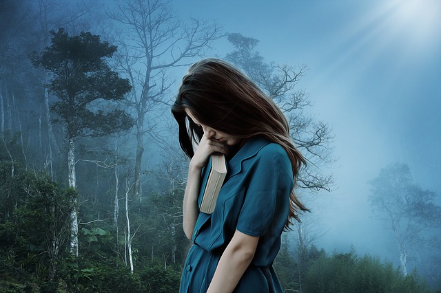 Woman looking downward in contemplation of possibly a recent tragedy with book in hand near face with bluish fog and trees in background