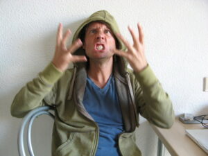 Angry man in a room at a desk wearing a green hoodie and a blue shirt shaking his hands in front of his face