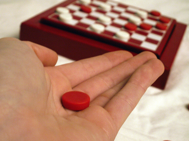 Hand holding a red checker piece with a checker game in the background