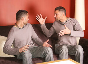 Photo of two identical men seated on couch in argument with one man's hands in the air