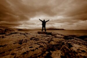 sepia photo of man with arms raised standing on rocky ground facing towards large body of water on cloudy day