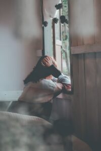 woman sitting on edge of bed with head in hands looking down out open window