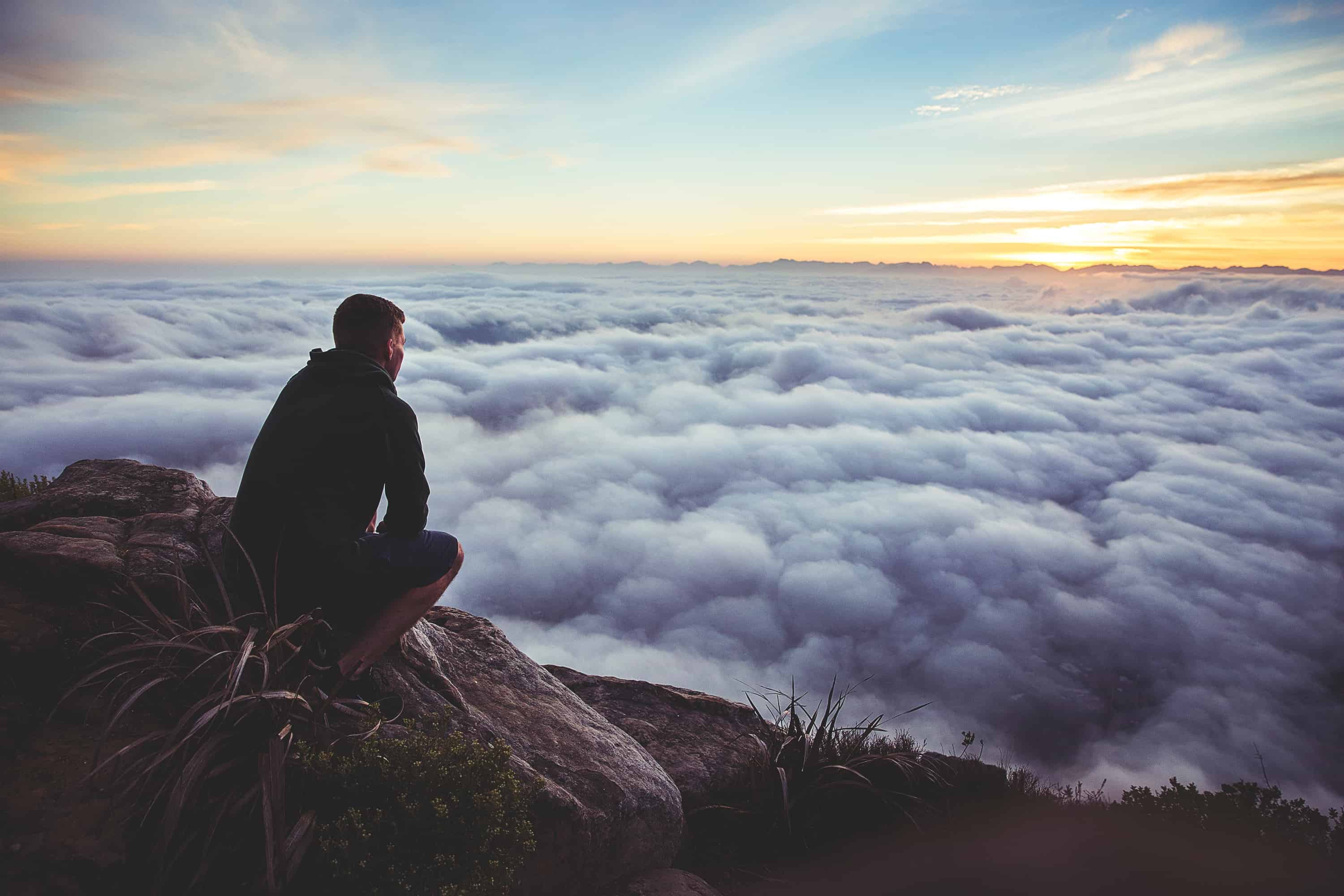 man sitting on mountainside overlooking clouds below
