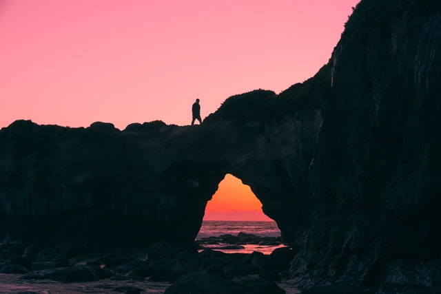 A photograph of a man walking on a natural rock arch formation over water with the pink sky of a sunset in the background