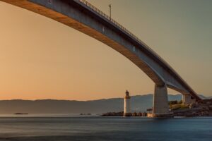 A tall modern bridge over water with lighthouse at end and mountains in background