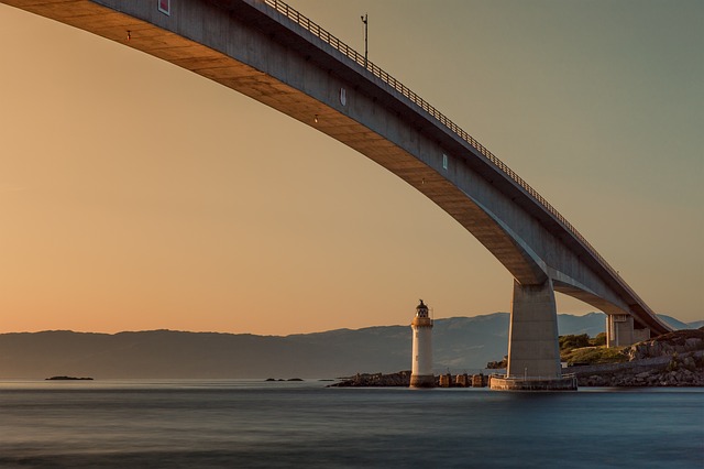 A tall modern bridge over water with lighthouse at end and mountains in background
