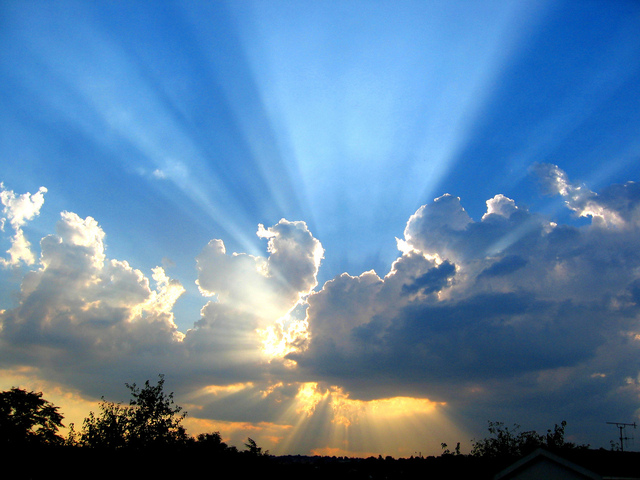 A photo of the sun behind a cluster of clouds with beams streaming from the top and orange sky beneath over a hilltop with some shrubbery
