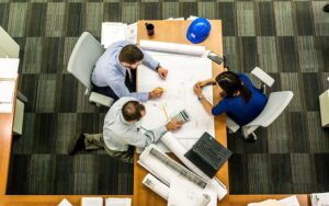 overhead photo of three people in meeting at table containg blueprint