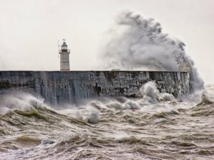 A giant wave crashing near a small lighthouse on a large stone foundation in the midst of very rough water.