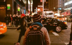 The backside of a person wearing a backpack and a knit hat attempting to cross a busy street at night in a bustling well lit downtown