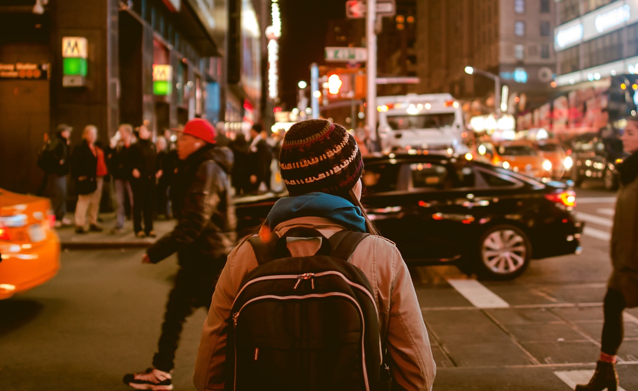 The backside of a person wearing a backpack and a knit hat attempting to cross a busy street at night in a bustling well lit downtown