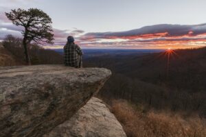 A person in a plaid shirt sitting on a cliff overlooking a dark valley with the sun rising in the background