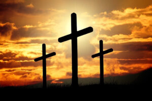 Silhouette of three crosses on hillside with orange sky containing clouds and sun in the background