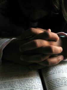 Hands folded in prayer on an open Bible under soft incandescent light