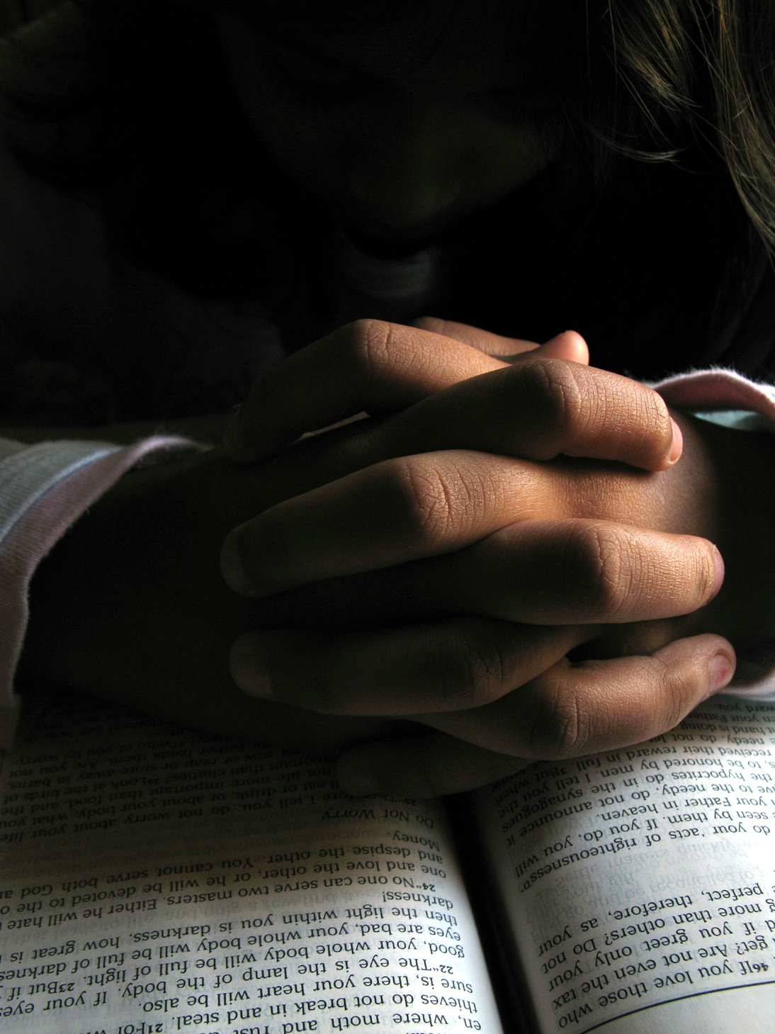 Hands folded in prayer on an open Bible under soft incandescent light