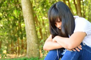 Girl with black hair, white shirt and blue jeans leaning over on knees while seated with serious or sad expression on her face