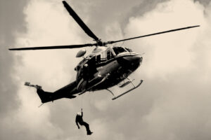 A sepia tinted black and white picture of a man being lowered from a helicopter hovering in the sky with clouds in the background