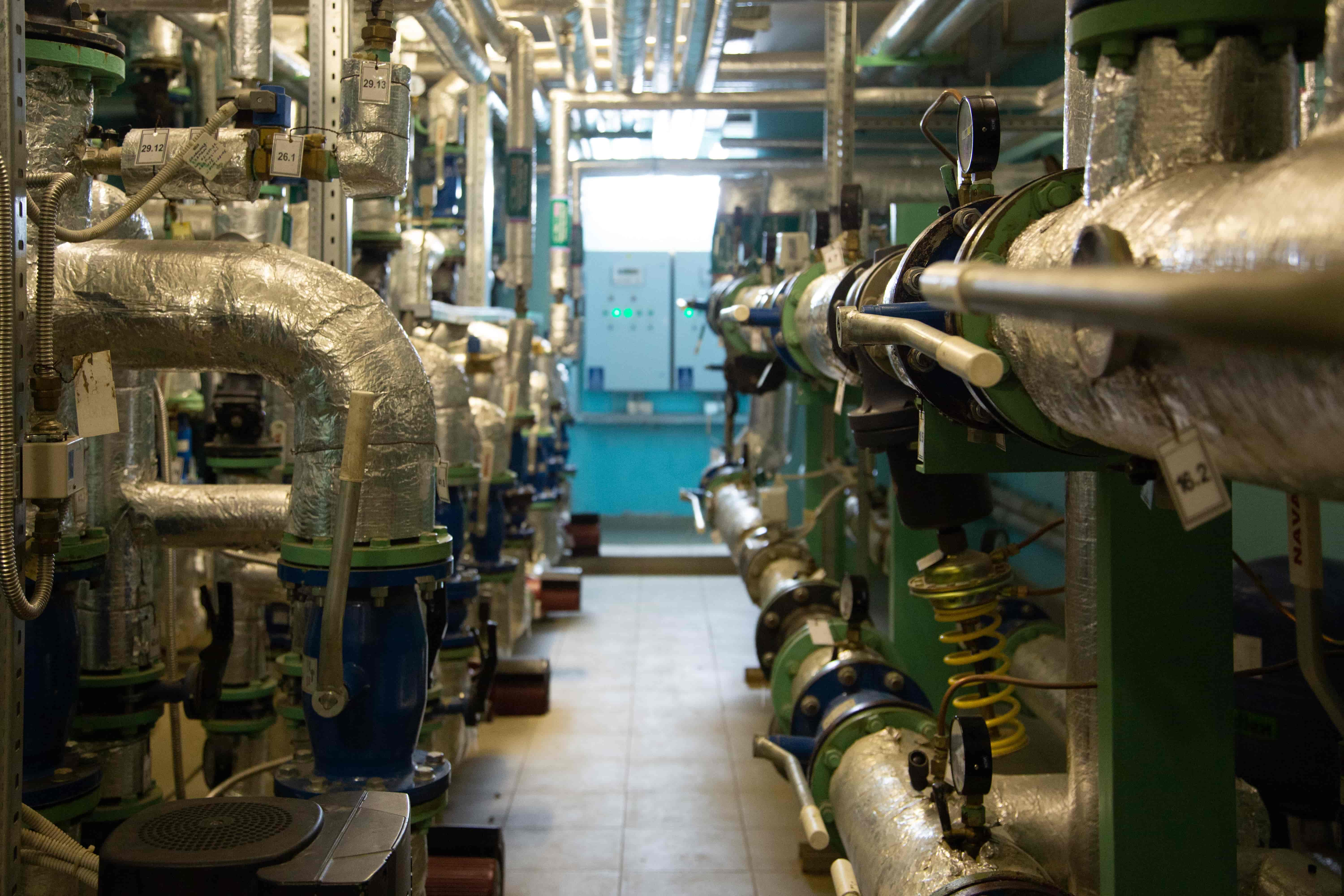A narrow walkway with various insulated pipes of industrial equipment and control panels at the end of the walk