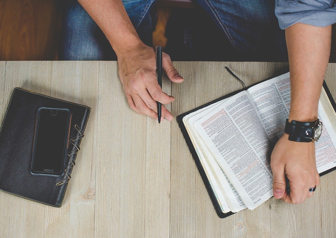 man sitting a a desk with left hand holding an open Bible and the right hand holding a pen near a book and phone