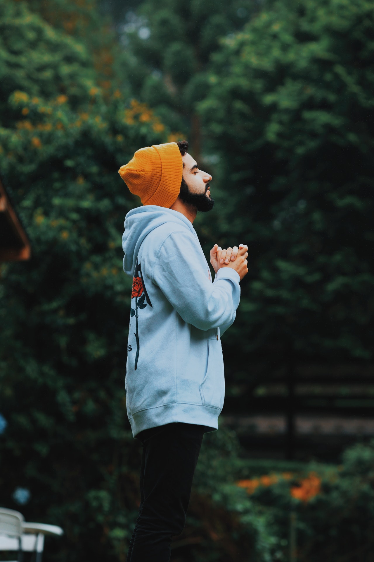A man with a beard standing and wearing a dark orange knit hat, a light blue hooded sweatshirt and dark blue pants with eyes closed and hands held together in prayer with trees in the background.
