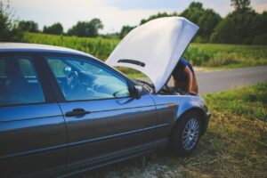 A young man bent over the engine of a car with hood open alongside a small rural road