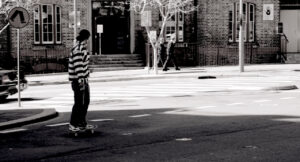 A black and white photo of a young man riding a skateboard into small intersection of a city with a building and two men walking in the background