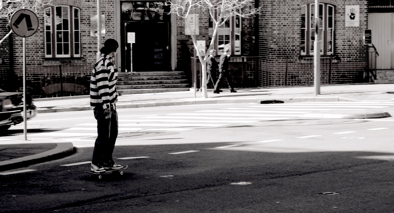 A black and white photo of a young man riding a skateboard into small intersection of a city with a building and two men walking in the background