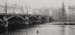 Black and white image of a multi-arched stone bridge over a river on a cloudy, snowy day in a city