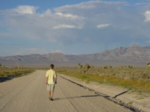 A man in tan shorts and yellow shirt walking on dirt road in an arrid region with mountains, clouds and blue sky in the background