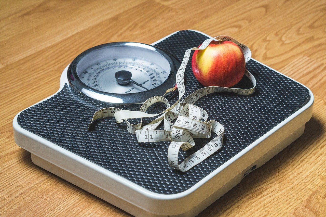A scale resting on a wooden floor with a tape measure and piece of fruit.