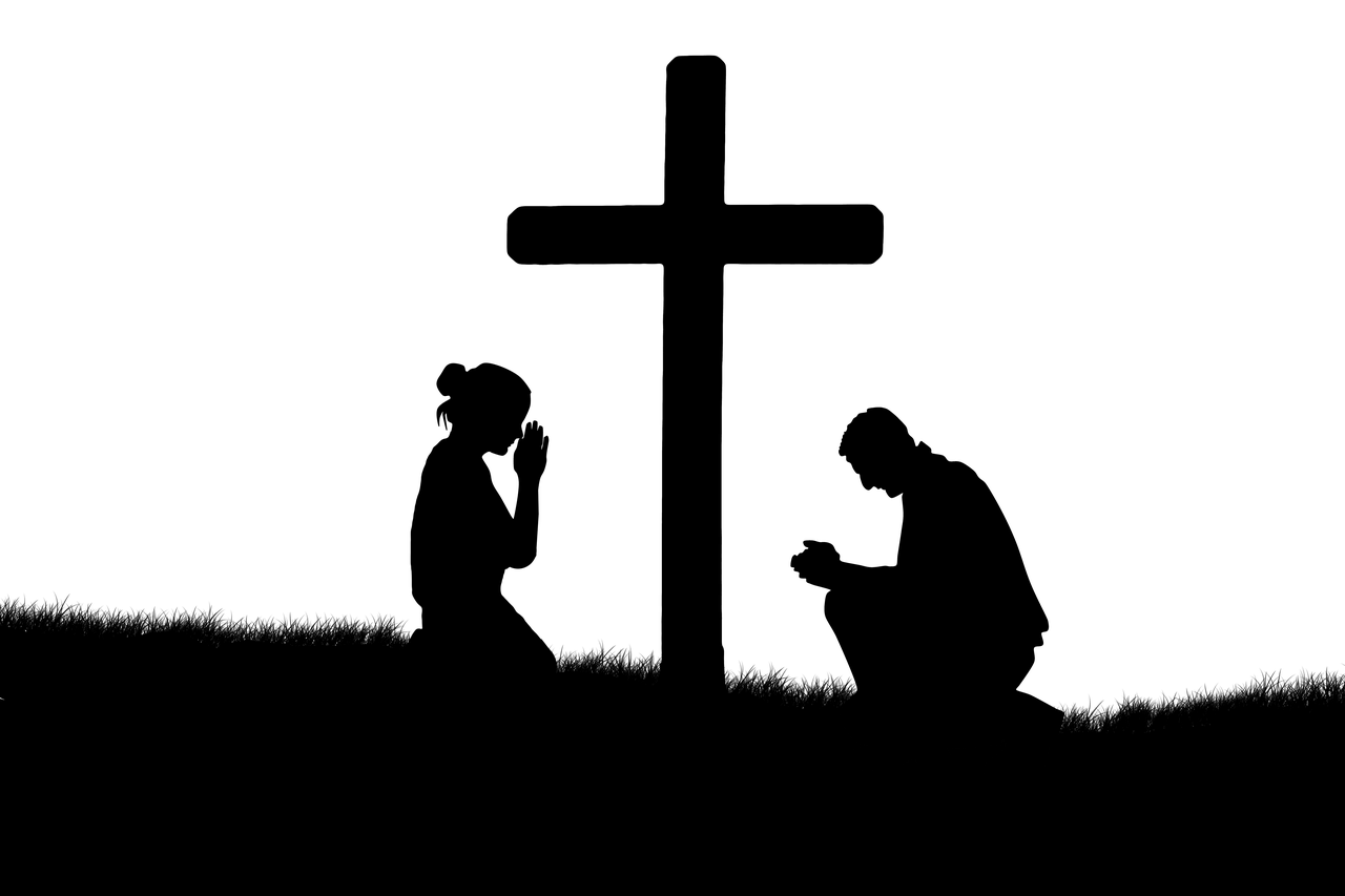 A silhouette of a woman and a man kneeling with arms together in prayer before a cross