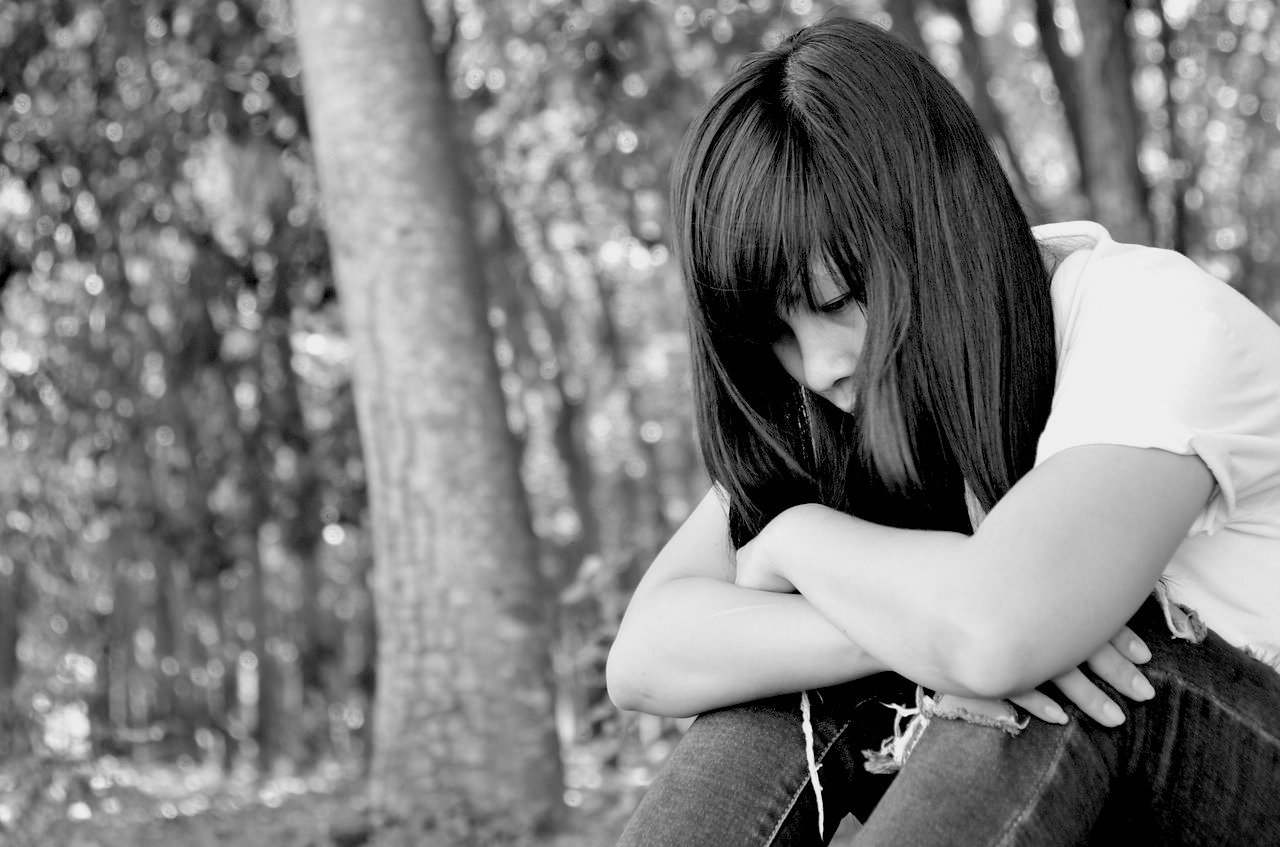 Girl with black hair, white shirt and blue jeans leaning over on knees while seated with serious or sad expression on her face