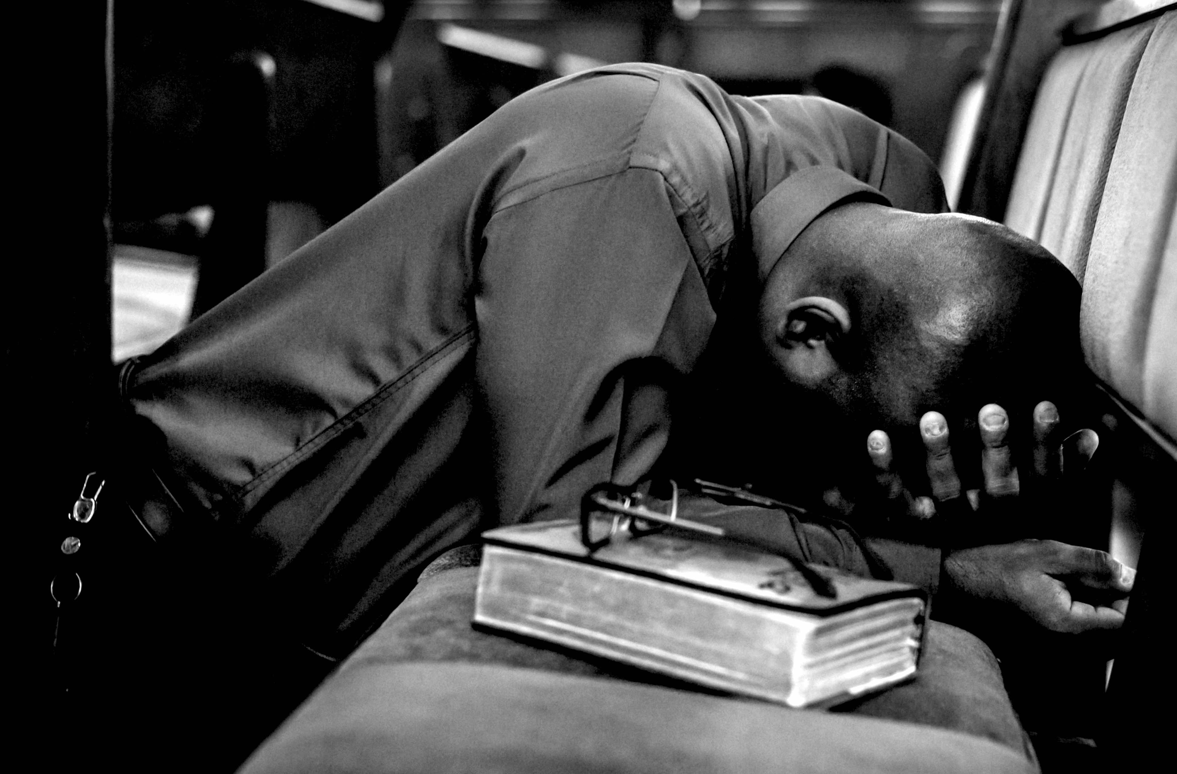 A black and white picture of a man in prayer at a church pew with bible and glasses nearby