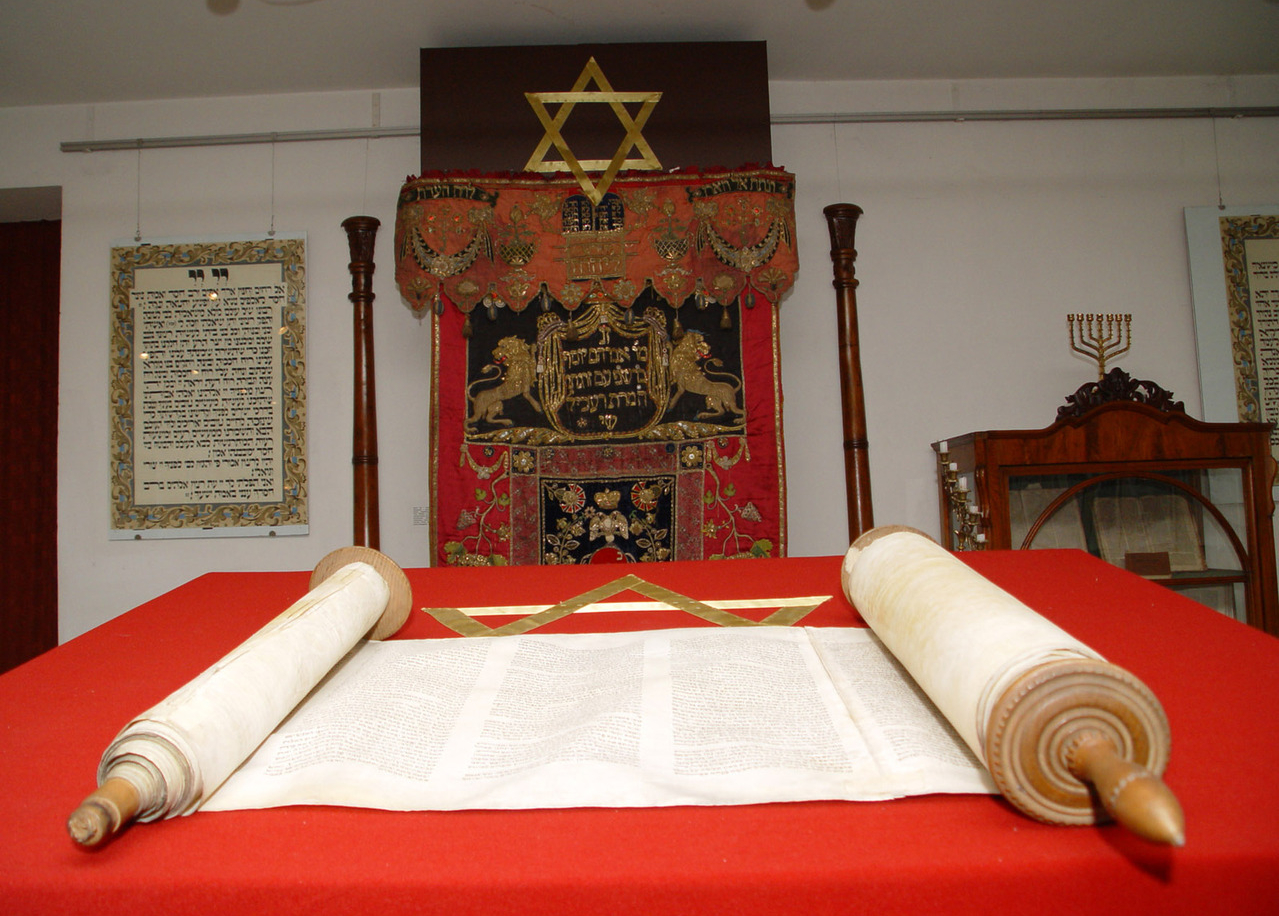 A scroll of the Torah opened on a red table with various Jewish decorum in the background