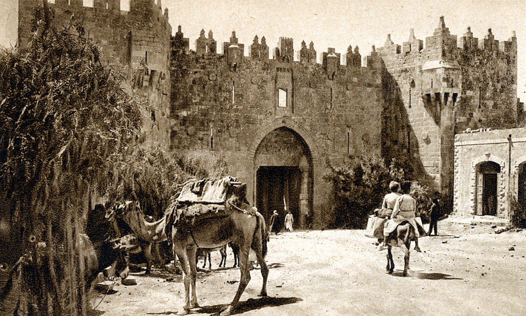 A sepia postcard from the 1920s of the Damascus Gate in Jerusalem with camels and a person on a donkey