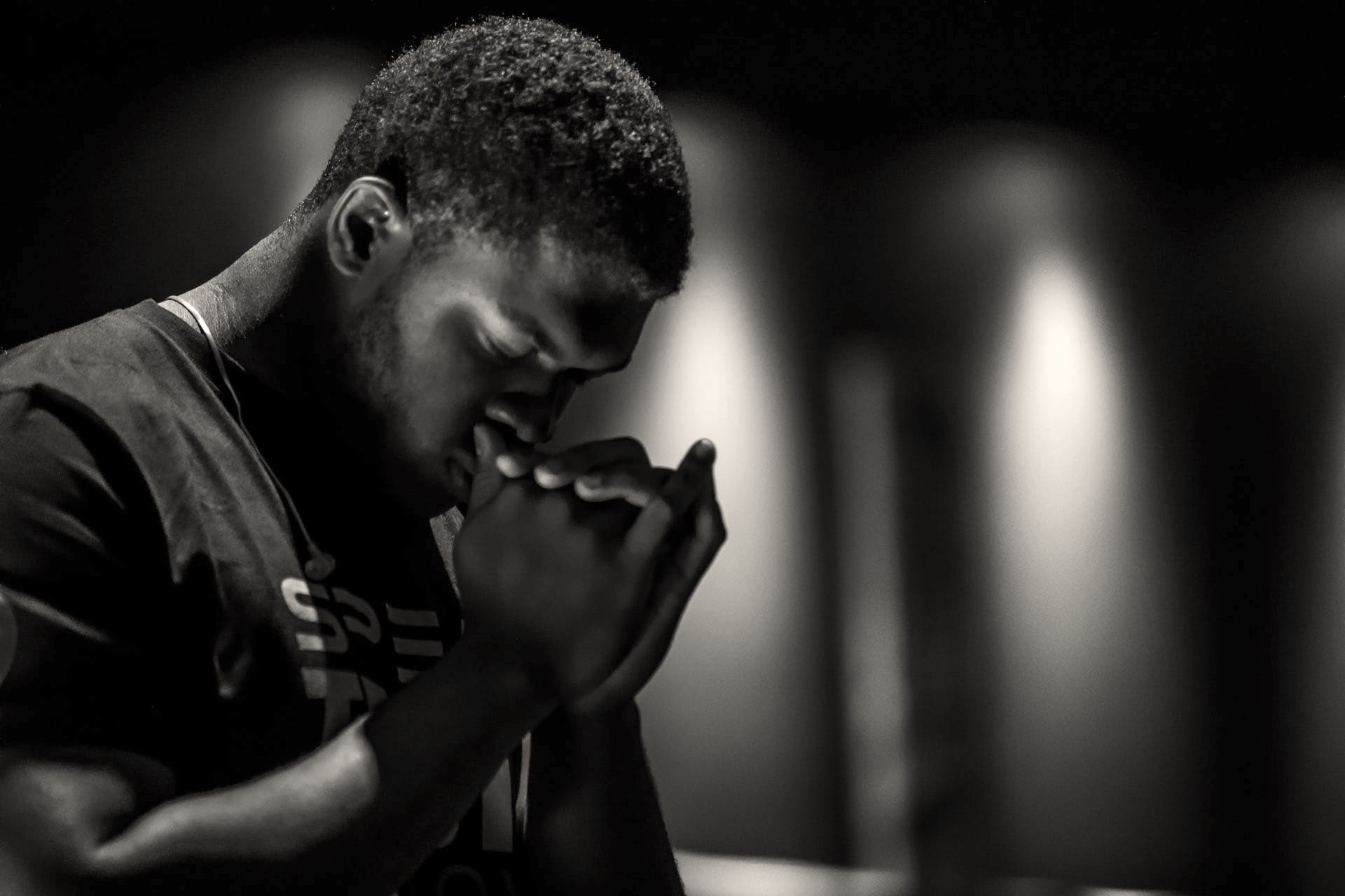 A black and white photo of a African American man bowing his head with folded hands in prayer