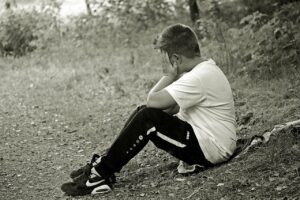 A black & white photo of the side view of a boy seated with his knees propped up and left hand pushed against the side of his face that is looking away.