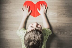 Overhead view of boy in green shirt laying stomach down on floor with hands resting on cut-out of a red paper heart