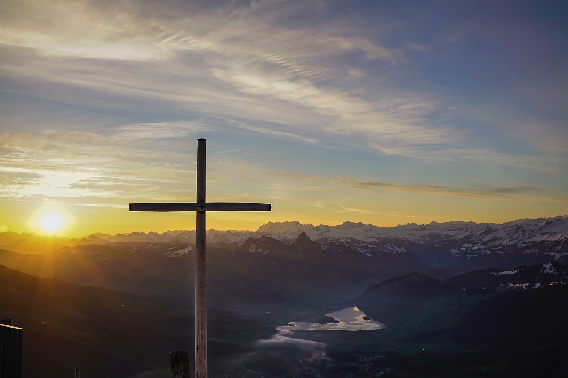 A cross on a hillside overlooking a large valley with mountains and a meandering river in the distance with the sun rising in the background