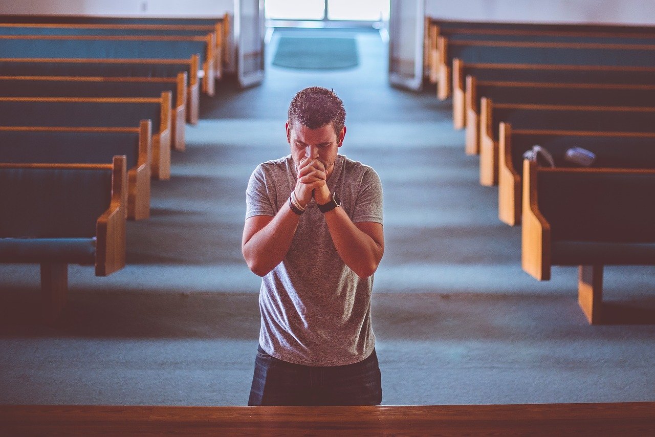 The front view of a man standing at the altar of a small church with his eyes closed and his hands raised and folded in prayer near his mouth.