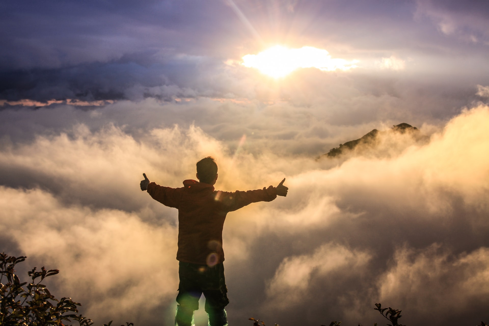 A man standing above the clouds with his arms outstretched and thumbs up with clouds and sun in the distance.