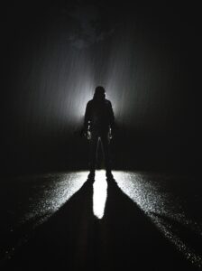 A black and white silhouette of a person in the darkness with a light behind them and rain falling